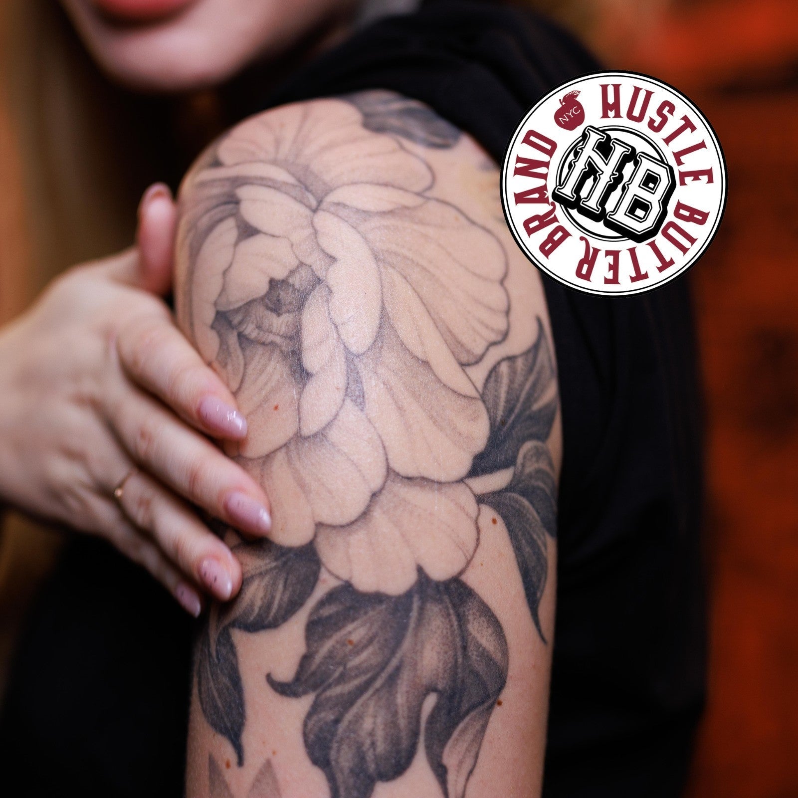 A fine-line black and grey floral tattoo on a woman's upper arm, with the Hustle Butter brand stamp in the top right corner.